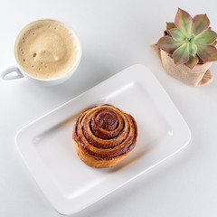 Cup of coffee, cinnamon roll bun pastry and small succulent plant over the white background.