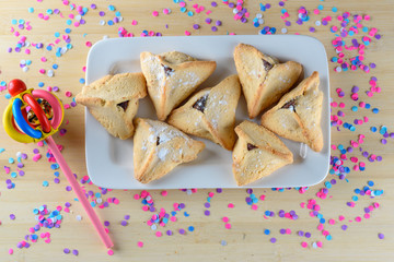 Hamantaschen cookies on white dish and wooden noisemaker or gragger on wood table. Colorful confetti background. Purim holiday celebration. Top view.