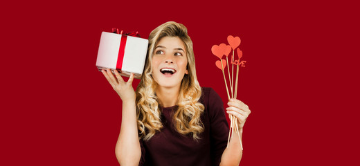 Young beautiful girl with a white gift with a red bow and hearts in her hands rejoices on an isolated red background. Valentine day concept.