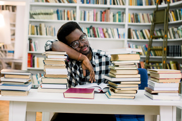 Ethnic african american guy lying at table surrounded by books in library. Student is bored and tired.