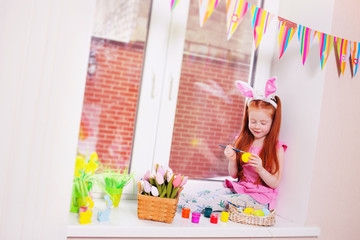 a small pretty girl with red hair with rabbit ears on her head is painting eggs for an Easter basket on the windowsill against the window and Easter decor. 