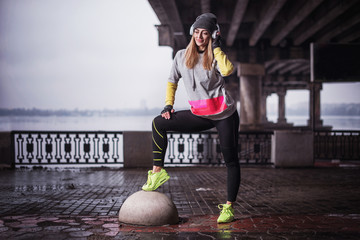 . sporty woman in headphones runner stretching legs before run under the bridge. Young woman resting after running in winter