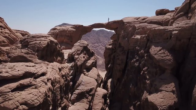 Drone flying under natural arch stone bridge while hiker is climbing across in desert