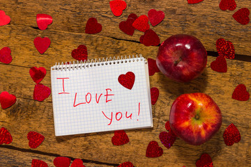 On a wooden rustic table are two red apples, hearts and a notebook with the inscription I LOVE you Valentine's Day