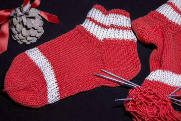 Red socks, needles and a ball on a black background. Next to a festive pine cone.