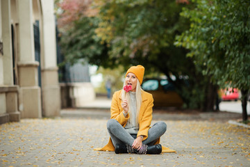 Saint Valentine's day concept. Fashion portrait blond young woman in yellow coat having fun with red lollipop heart over street background. 