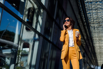 Beautiful business woman in yellow suit walking near business centre at the city, communicates via smartphone and drinks coffee. Business concept, communication, banker, manager and office worker.