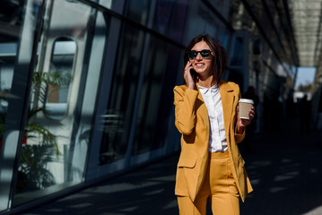 Obraz premium Smiling business woman in yellow suit walking near business centre at the city, communicates via smartphone and drinks coffee. Business concept, communication, banker, manager and office worker.