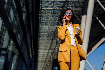 Obraz premium Young smiling business woman in sunglasses and yellow suit stands near office centre with cup of coffee and talks on smart phone. Lunch break among working day. Business lifestyle and success concept.