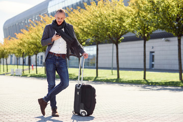 Young stylish man using his smart phone while standing with suitcase near exit the airport outdoors. Business style, traveler, modern lifestyle, business trip.