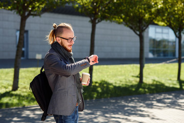 Fototapeta premium Young successful businessman in a grey jacket with scarf walks near the business centre, drinks coffee and looking on the watches. Concept of business, banker, manager.
