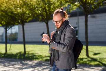 Fototapeta premium Young successful businessman in a grey jacket with scarf walks near the business centre, drinks coffee and looking on the watches. Concept of business, banker, manager.