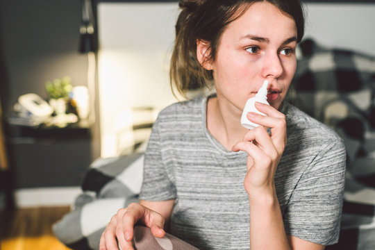 The theme is seasonal cold, runny nose flu virus infection. Young caucasian woman at home bedroom bed uses spray drops of medicine in her nose for snot. Allergic renitis and sinusitis