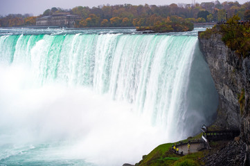 Beautiful view of the amazing Niagara Falls seen from the Canadian border in Autumn. Some tourists with a yellow raincoat are shooting selfies under the falls.