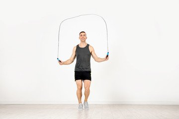 Full length portrait of young sportive man training with jump rope in light room