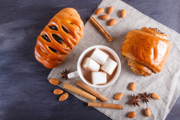 a cup of hot chocolate with marshmallow, buns, almonds and spices on black wooden background.