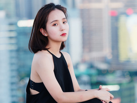 Beautiful young brunette woman in sexy black dress sitting on top of mansion roof with blur Shanghai Bund landmark buildings background with dusk light. Emotions, people, beauty and lifestyle concept.