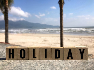Motivational and inspirational quote - ‘HOLIDAY’ written on wooden blocks. Blurred background of a beach.