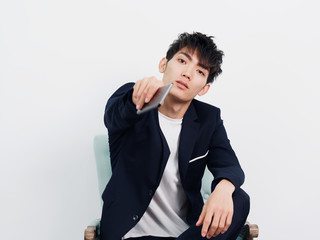 Portrait of handsome Chinese young man in dark blue leisure suit sitting in armchair and looks unhappy with mobile phone in hand pointed at you.