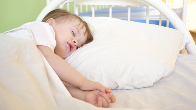 charming baby falls asleep on white bed in his bed in room at home. concept of sleeping child. child wants to sleep and rubs his eyes with his hands. kid sleeps in hospital ward. close-up