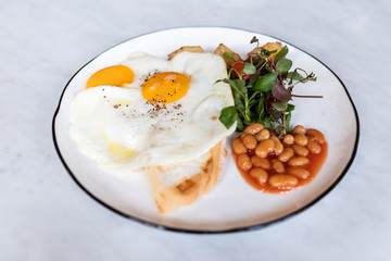 Delicious English breakfast with two fries eggs, beans, and salad on marble table.