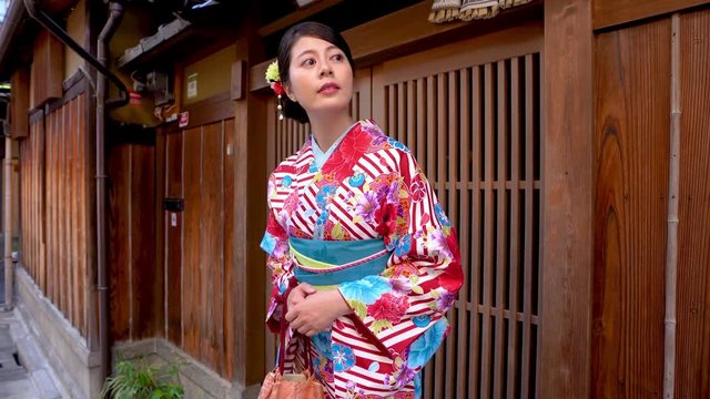 elegant local woman in flower pattern kimono standing in front of wooden home waiting for friends join festival together. young japanese lady in traditional costume looking around carrying purse.