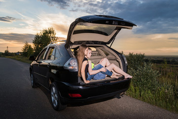 Two girls sitting in the trunk of a car. Friends relax in the summer evening and sunset behind them