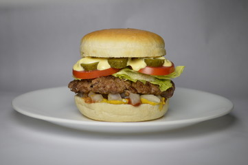 American burger - bread, meat, lettuce, tomato, pickles, onions whit light mayonnaise, over  a white plate- no background