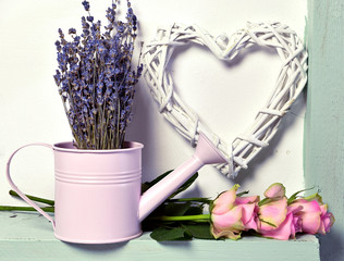Cute watering can with lavender bunch, rose flowers and heart symbol on white wall. Romantic vintage valentine’s day and wedding background
