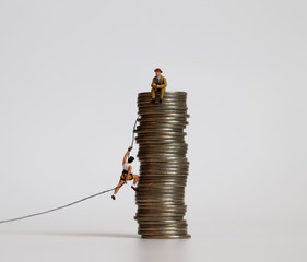 A miniature woman trying to climb with a rope on a pile of coins and miniature man sitting on a...