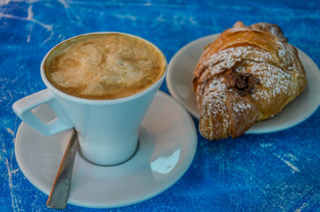 Cappuccino cup and saucer with croissant on blue background