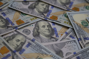 The face of Benjamin Franklin, shown on the front of the U.S. $100 bill, is shown among other loosely scattered bills of the same denomination in an angled view. This design was issued in 2013.