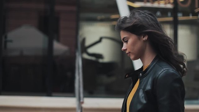 Pretty young woman in the leather jacket is walking near the business center. The girl fixes her hair looks around the place