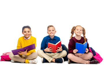 cheerful schoolchildren sitting, holding books and looking at camera isolated on white