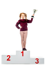 cheerful schoolgirl standing on winner podium, smiling, holding trophy cup and looking at camera...