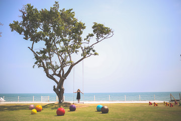 young girl swinging on a swing between ิbigtree on the beach of a tropical island. Young woman watching the ocean on a sunny day