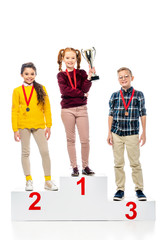 smiling preteen kids with medals and trophy cup standing on winner pedestal, smiling and looking at camera isolated on white