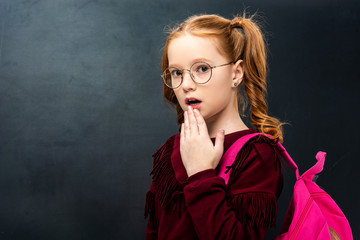 surprised schoolgirl in glasses with pink backpack looking at camera on black background