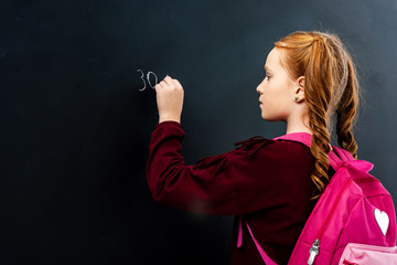 schoolgirl with pink backpack writing on blackboard with chalk
