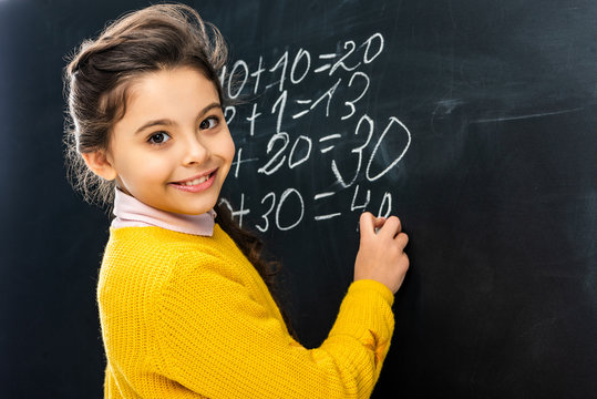 smiling schoolgirl writing on blackboard with chalk and looking at camera