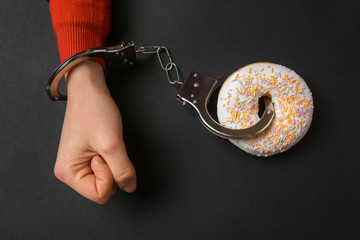 Woman handcuffed to tasty doughnut on dark background. Concept of addiction - obrazy, fototapety, plakaty