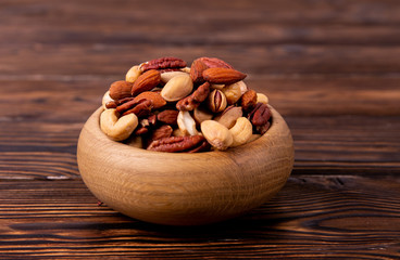 Mixed nuts in wooden bowl and scattered on table. Trail mix of pecan, almond, macadamia & brazil edible nuts with walnut hazelnut on wood textured surface. Background, copy space, top view, close up.