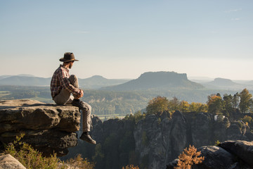 Young bearded male traveler in hat stand on the cliff in sunny day 