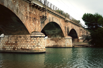 An old stone bridge over a river in Italy. Green running water, bricks, trees.

