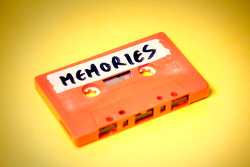 A vintage cassette tape (obsolete music technology), orange on a yellow surface, angled shot, carrying a label with the handwritten text Memories.
