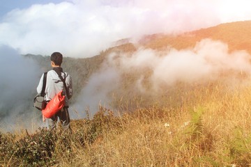 Asian man standing, carrying red bag and looking to white mist and mountain view and white clouds with orange sunlight flare at Doi Inthanon National  Park, Chiangmai, Thailand - Beauty of Nature