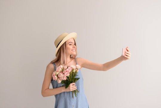 Beautiful young woman with bouquet of flowers taking selfie on grey background