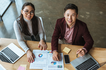 high angle view of happy young asian business colleagues sitting together and smiling at camera in...