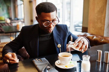 Fashionable african american man in suit and glasses pours cream into coffee at cafe.