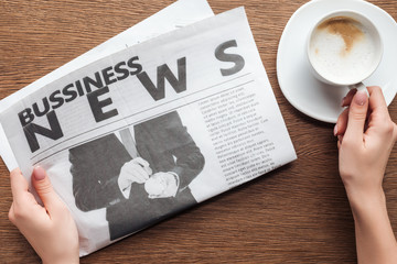 cropped image of journalist holding business newspaper and cup of coffee at wooden table
